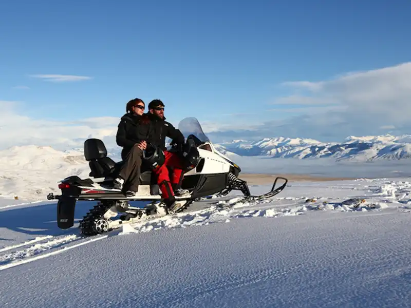 Pareja en moto de nieve biplaza con las montañas nevadas de la Cordillera de los Andes de fondo, travesía invernal en El Calafate.